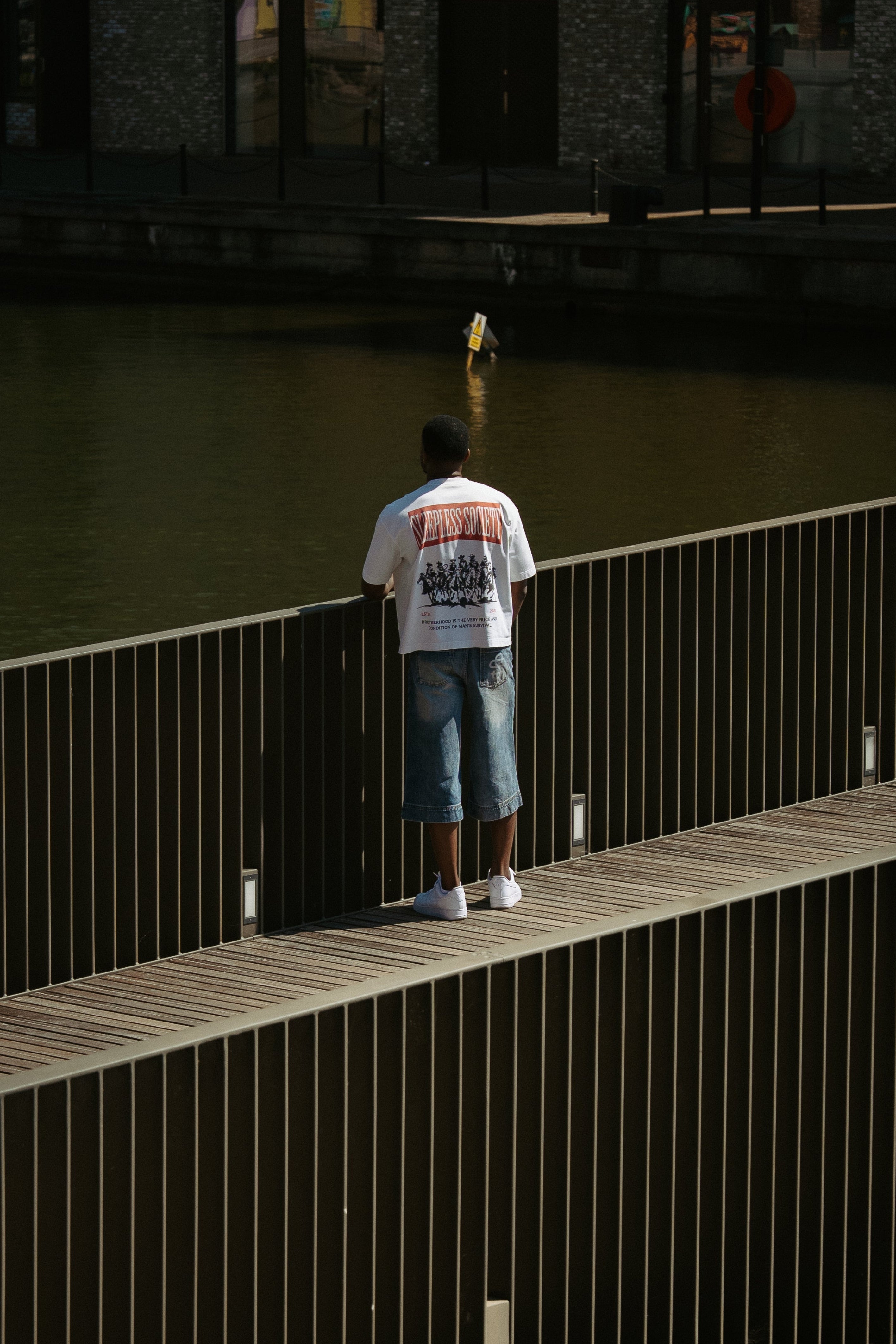 A man stands on a wooden boardwalk overlooking a body of water, wearing a white graphic t-shirt and denim shorts.